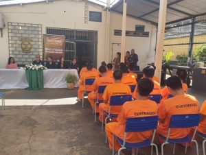 internos enfileirados, vestidos com roupas laranja, em cadeiras e a frente mesa com autoridades